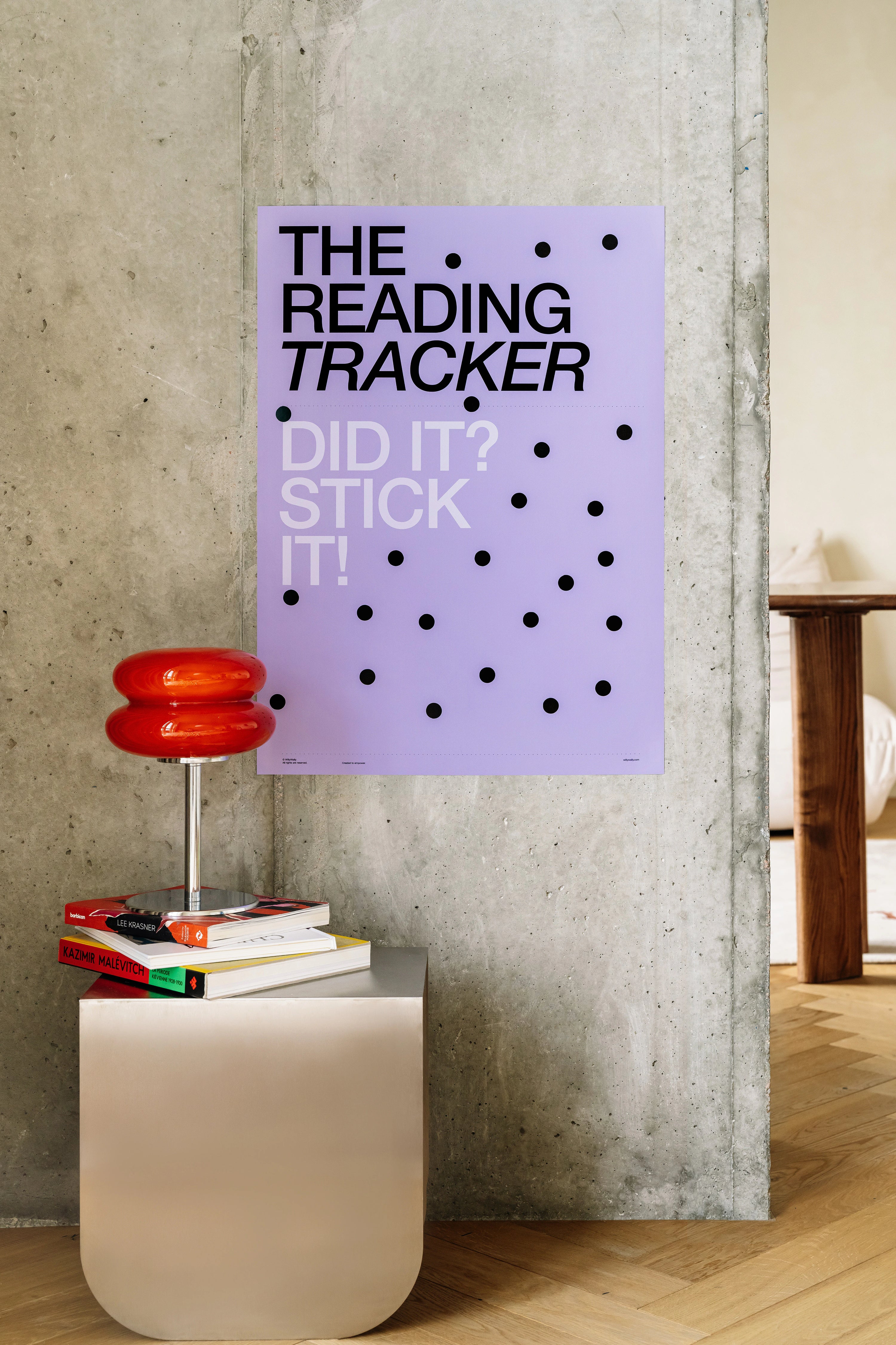 Purple poster with text 'The Reading Tracker' on a concrete wall, with books and a red object on a table below.