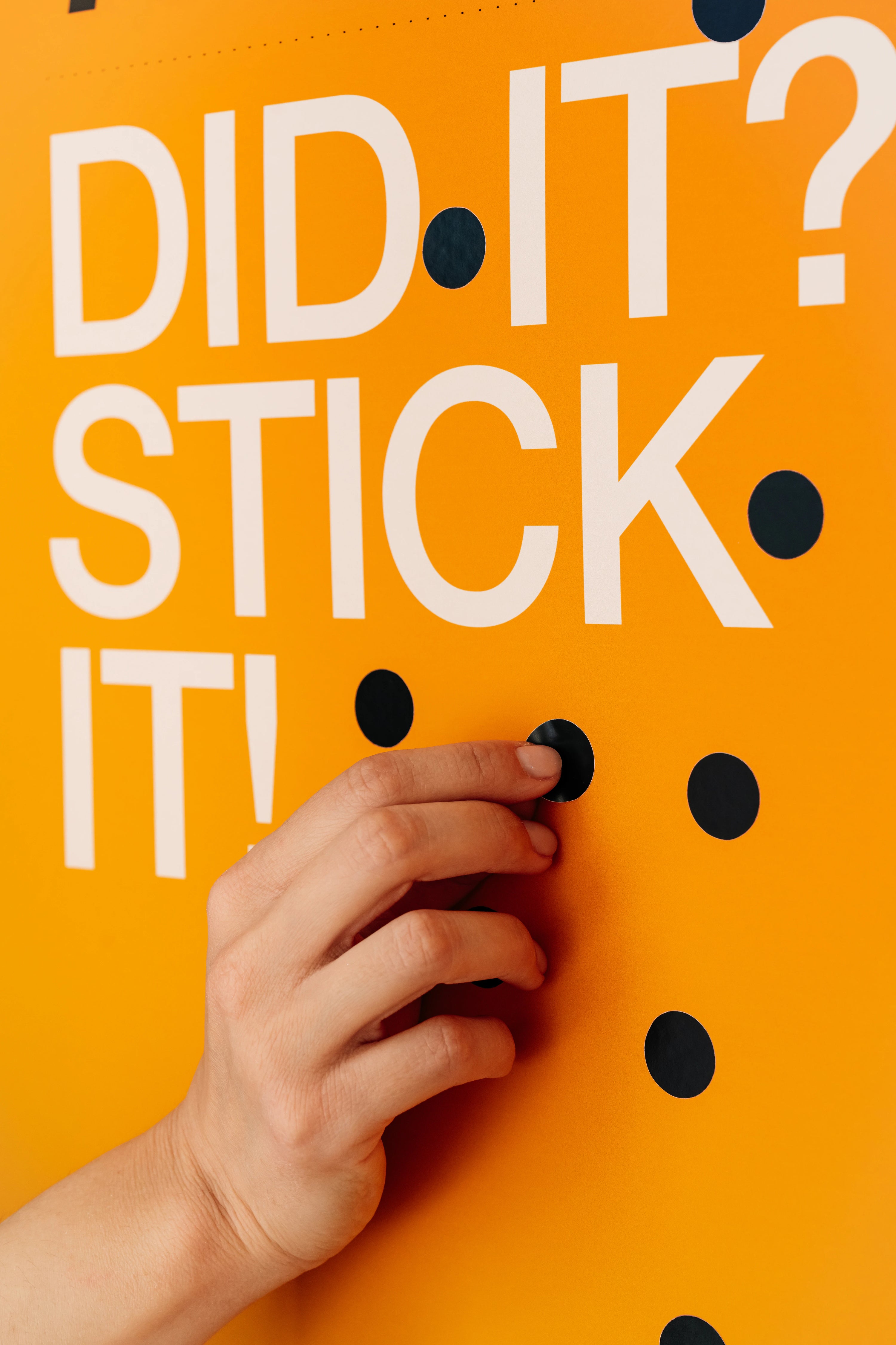 Hand pressing a black dot onto an orange surface with text 'DID IT? STICK IT!'